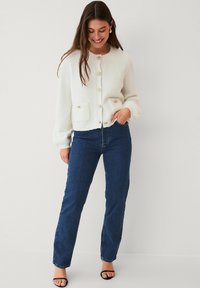 Witte gebreide cardigan met ronde hals, knoopsluiting en twee voorzakken. Gecombineerd met blauwe rechtlopende jeans en zwarte sandalen.