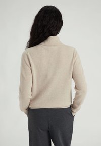 Maglione lavorato a maglia beige con colletto alto e texture a coste, abbinato a pantaloni grigi scuri. Il maglione ha una vita aderente e maniche lunghe.