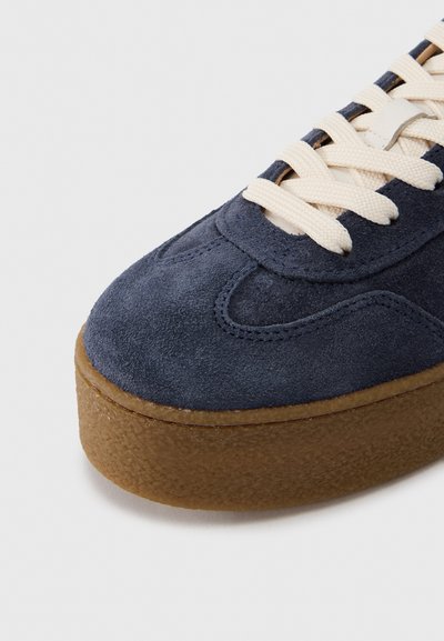Σκούρο μπλε σουέντ αθλητικό παπούτσι με εκρού κορδόνια, ανοιχτό καφέ υφασμένο πέλμα, στρογγυλεμένη μύτη και ραμμένες λεπτομέρειες κατά μήκος του πάνω μέρους.