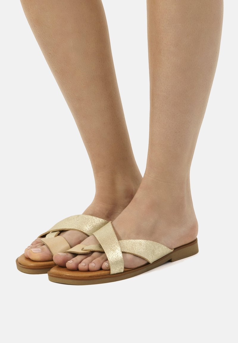Sandali piatti metallici oro con cinturini incrociati, suola sintetica marrone. Presentano un design open toe per una maggiore traspirabilità.