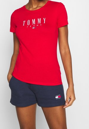 Personne portant un t-shirt rouge à manches courtes avec le texte "TOMMY JEANS" et un short bleu marine avec un écusson logo assorti, se tenant devant un fond blanc.