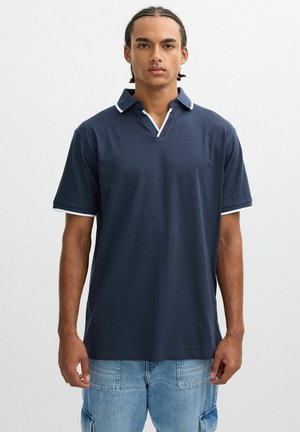 Joven con cabello trenzado que lleva una camisa polo azul marino con ribete blanco y pantalones cargo de mezclilla azul claro, de pie mirando hacia adelante.