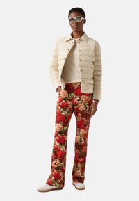 Kremowa kurtka z podszewką, beżowy top, czerwone spodnie w kwiaty, białe trampki. Dodatki obejmują okrągłe okulary przeciwsłoneczne. Strój wyróżnia się wyrazistymi kolorami i wzorami.