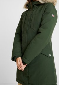 Grüne gefütterte Parka mit fellgesäumter Kapuze, Frontreißverschluss und Seitentaschen. Verfügt über eine Brusttasche, elastische Bündchen und Logodetail am Ärmel.