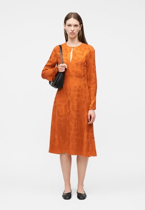 Femme portant une robe midi orange à manches longues avec des motifs subtils, des ballerines noires, et tenant un petit sac à bandoulière noir.