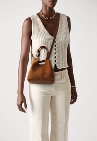 Brown suede handbag with a structured shape, featuring a short handle and adjustable strap. Worn with a knitted cream top and light pants.