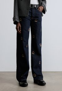 Tume sinised laiade säärtega teksapüksid, millel on tikandid kroonidest punases ja kuldsetes toonides. Valmistatud siledapinnalisest teksariidest. Sobitatud mustade jalanõudega.