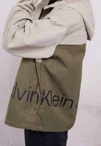 Osoba ubrana w dwukolorową kurtkę z kapturem z oliwkowymi i beżowymi panelami oraz dużym czarnym logo Calvin Klein na plecach.