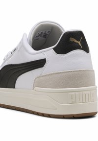 Baskets PUMA avec un dessus en cuir blanc, une bande noire, un talon texturé beige et une semelle en caoutchouc avec un logo en relief.