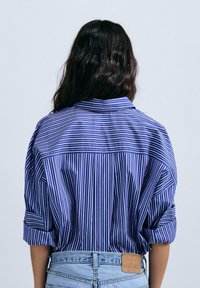 Cămașă oversized cu dungi albastre și albe, cu mâneci răsucite și detalii la spate, realizată dintr-un material ușor. Asortată cu pantaloni șort din denim.