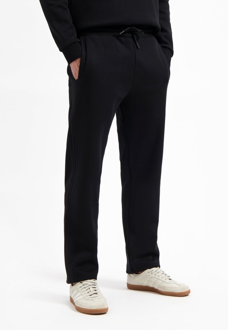 Schwarze Sweatpants aus weichem Material, mit seitlichen Taschen und einem elastischen Bund mit Kordelzug. Kombiniert mit hellen Jogging-Schuhen.