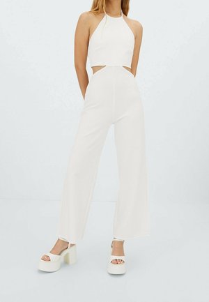 Woman wearing white sleeveless halter jumpsuit with side cutouts and wide-leg pants, paired with white platform sandals.