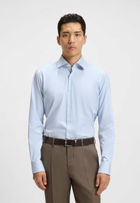 Camicia da uomo a maniche lunghe blu chiaro con sottili righe verticali, dotata di colletto a punta e chiusura frontale con bottoni, abbinata a pantaloni marroni.