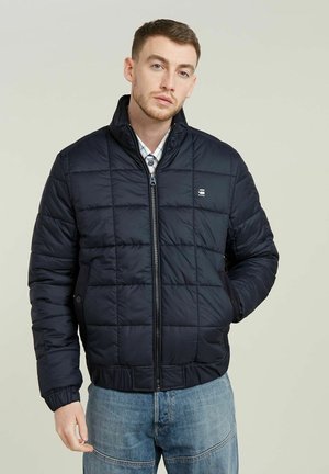 Navyblaue Pufferjacke mit einem gesteppten Design, hohem Kragen, vorderem Reißverschluss und Seitentaschen. Darunter wird ein kariertes Hemd getragen.