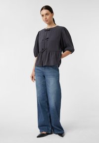 Bluse in Anthrazit mit Puffärmeln, Schleifen vorne und einem Peplum-Saum, kombiniert mit weitem, blauen Jeans und spitzen, schwarzen Ballerinas.