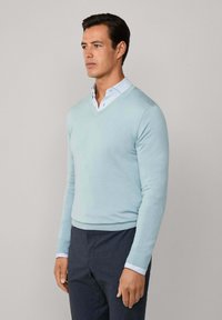 Svetlo modrý sveter s výstrihom do V, vyrobený z jemného pleteného materiálu, s dlhými rukávmi a priliehavým strihom, prekrytý košeľou s golierom.