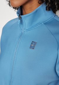 Hellblauer Reißverschlussjacke mit hohem Kragen, glatter Textur und kleinem gesticktem Logo auf der Brust.