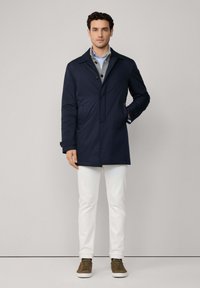 Marineblauwe, middellange jas van waterafstotende stof met een knoopsluiting, zijzakken en een ingesneden kraag, gedragen over een grijze trui.