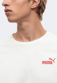 Νεαρό άτομο που φοράει λευκό μπλουζάκι Puma με κόκκινο λογότυπο Puma στο στήθος, κοιτάζοντας προς τα αριστερά σε απλό φόντο.