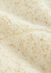 Cremfarbende Fleece-Decke mit einer weichen, strukturierten Oberfläche. Verfügt über kleine braune Flecken, die ein gemütliches Erscheinungsbild bieten.