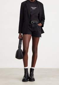 Person som bär en svart blazer, svart croppad topp från Calvin Klein Jeans, svarta denimshorts med bälte, svarta stövlar, vita strumpor och håller en svart handväska.