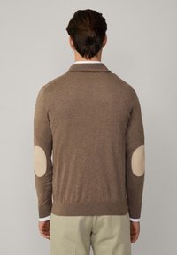 Smeđi kardigan s ovratnikom, s laganim bež zakrpama na laktovima i prilagođenim dizajnom. Izrađen od mekanog materijala, ima glatku teksturu.