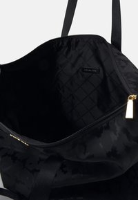 Musta liikuntakassi, jonka vetoketju on auki ja jonka sisälle näkyy brändätty vuori ja kultainen vetoketjun vetopidike. Kaksi mustaa remmiä näkyy valkoista taustaa vasten.