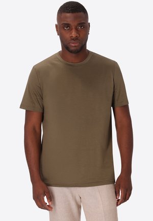 Uomo che indossa una maglietta verde oliva a maniche corte e pantaloni beige chiaro su uno sfondo bianco uniforme.