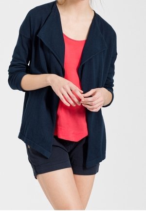 Femme portant un cardigan bleu marine ouvert sur un débardeur corail et un short bleu marine, les mains légèrement jointes au niveau de la taille.