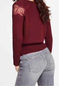 Burgundy neule, jossa on pitsiyksityiskohtia olkapäillä, ribbineuleiset hihansuut ja helma. Yhdistetty vaaleanharmaisiin farkkuihin, joissa on metallisia yksityiskohtia taskuissa.