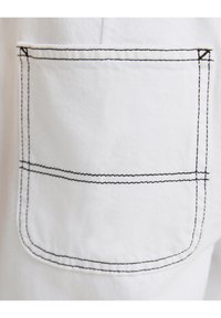 Bolsillo de tela blanca con detalles de costura negra, que presenta un borde superior redondeado y líneas rectas, indicativas de un diseño estructurado.