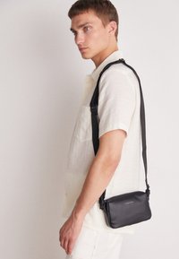 Bolso cruzado de cuero negro con una textura lisa, que presenta un cierre con cremallera y una correa ajustable, llevado sobre una camisa de color claro.