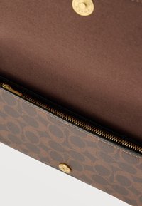 Καφέ πορτοφόλι σχεδιαστή με κυκλικό μοτίβο λογότυπου, χρυσό φερμουάρ και κλείσιμο με κουμπί, μερικώς ανοιχτό που δείχνει ομαλή εσωτερική επένδυση.