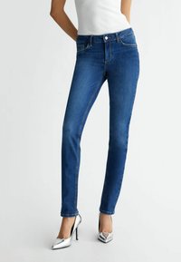 Jeans skinny blu scuro con una texture liscia. Presentano tasche frontali, chiusura con bottone e cuciture a contrasto. Indossati con tacchi alti argentati.