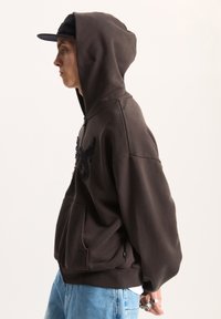 Sudadera con capucha marrón y bolsillo frontal, con un diseño bordado en negro, combinada con unos vaqueros de color azul claro. Textura suave, ajuste relajado.