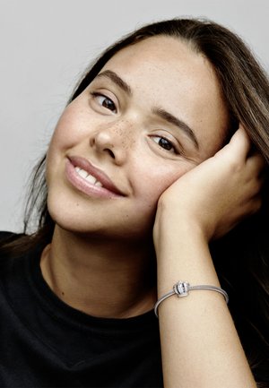 Silberarmband mit krönchenförmigem Verschluss, das über filigrane Details und ein glattes Texturband verfügt, getragen an einer Hand, die auf einem Gesicht ruht.
