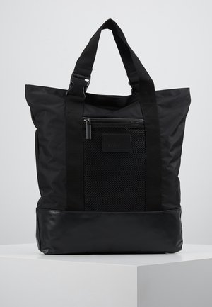 Sac fourre-tout noir avec poche avant en maille et doubles poignées courtes, fabriqué en tissu mixte avec fermeture éclair, posé sur un socle blanc.