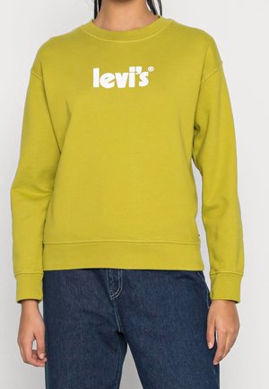 Osoba nosząca limonkowo zieloną bluzę Levi's z białym logo i ciemnoniebieskie dżinsy, stojąca z luźno opuszczonymi rękami wzdłuż ciała.