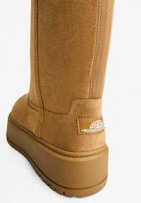Støvle lavet af beige ruskind med en platformsål, der har syninger og et logo-patch. Glat tekstur og design med rund tå.