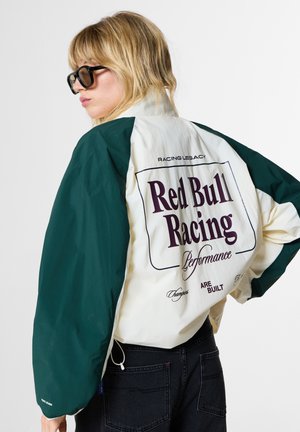 Vrouw met blond haar en zonnebril draagt een wit-groen jack met grote "Red Bull Racing Performance" tekst op de rug.