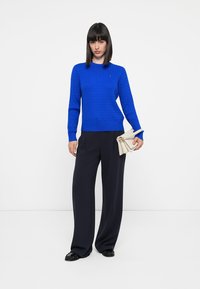 Pull texturé bleu cobalt avec un col rond, associé à un pantalon large noir et un sac à main beige. Le modèle se tient devant un fond blanc.