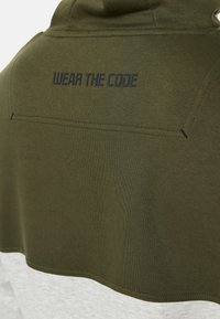 Olivengrøn hættebluse med en hvid nederste del. Har "WEAR THE CODE" i sort tekst på ryggen og synlige syninger.