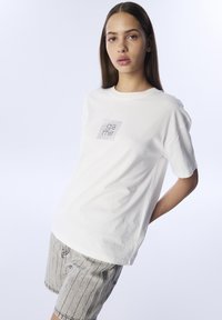 Camiseta de algodón blanca con cuello redondo y mangas cortas. Presenta un logo impreso con un diseño de rejilla en la parte frontal.