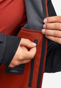 Czerwono-szara polarowa kurtka z zamykaną na zamek kieszenią. Posiada teksturowane wnętrze i kontrastowy czarny zamek, ręka angażująca się w uchwyt zamka.