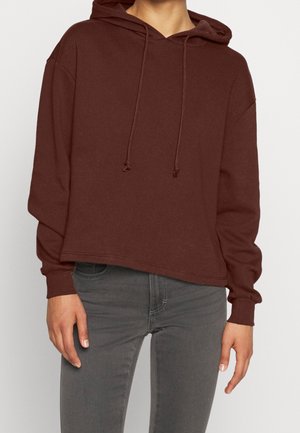 Persoon draagt een loszittende bruine hoodie met trekkoord en omgeslagen mouwen, gecombineerd met grijze slim-fit jeans.