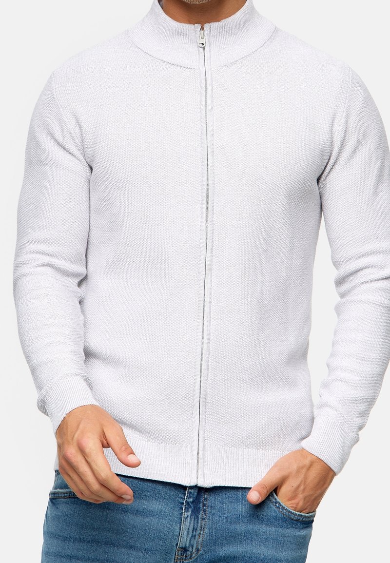 Homme portant un pull zippé blanc texturé et un jean bleu, une main dans la poche, sur un fond clair uni.