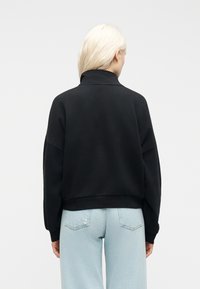 Γυναίκα με μακριά ξανθά μαλλιά που φοράει ένα μαύρο φούτερ με ψηλό γιακά και ανοιχτόχρωμο τζιν, εμφανιζόμενη από πίσω με φόντο μια απλή επιφάνεια.