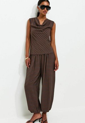 Vrouw die een bruin-zwart geruit mouwloos topje en bijpassende wijde broek draagt, oversized zwarte zonnebril, gouden creolen en zwarte hakken.