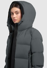 Chaqueta tipo plumón en gris oscuro con una gran capucha, tejido texturizado, cremallera frontal y ribete negro en contraste. Cuenta con bolsillos con cremallera en los laterales.