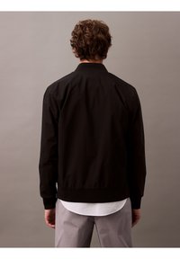 Schwarze Bomberjacke mit hohem Kragen, gerippten Bündchen und Bund. Glatte Textur, leichter Stoff, getragen über einem weißen Hemd.